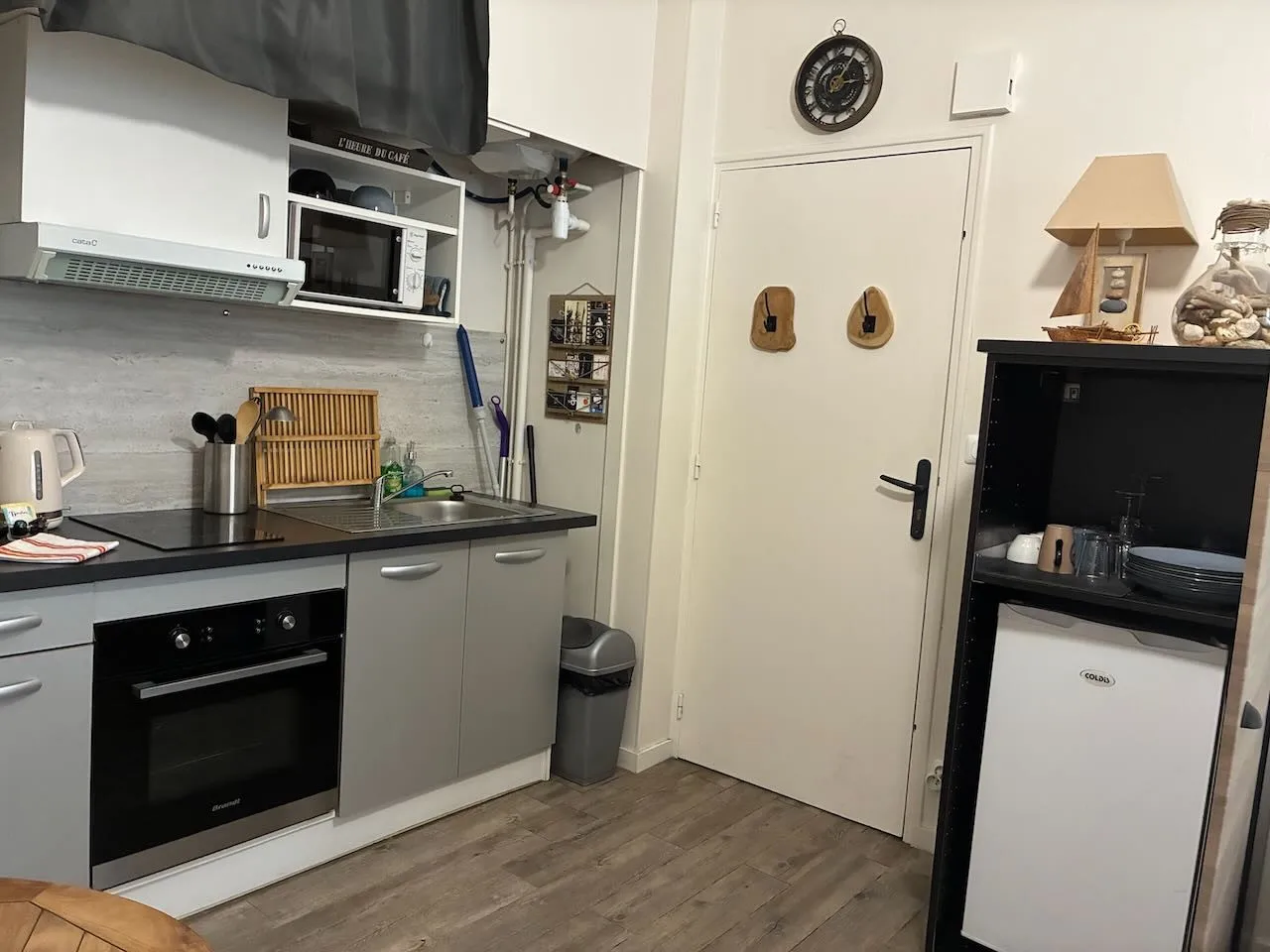Le coin cuisine, la porte d’entrée avec ses patères et l’armoire qui abrite le réfrigérateur